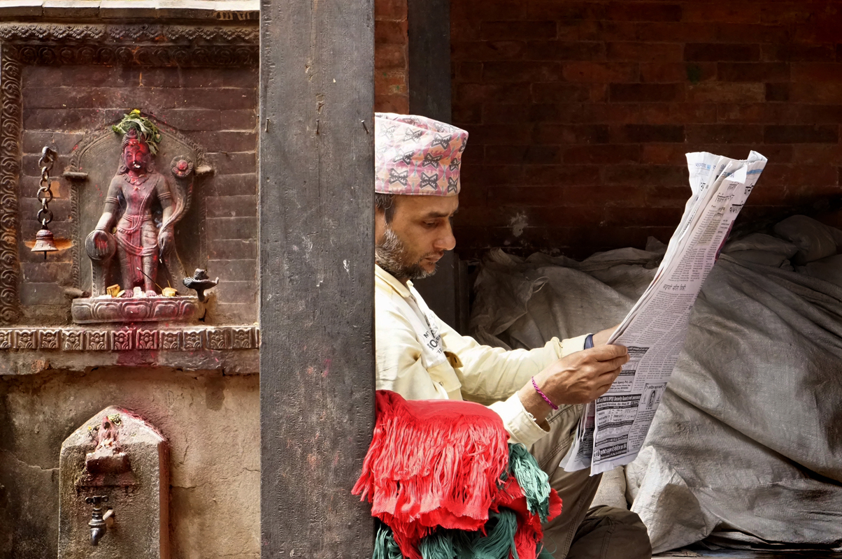 Street photography, Nepalese mens’ hat, man read newspaper, roadside shrine, religious effigy, stone carving, bell, incense, Bhaktapur, Nepal
