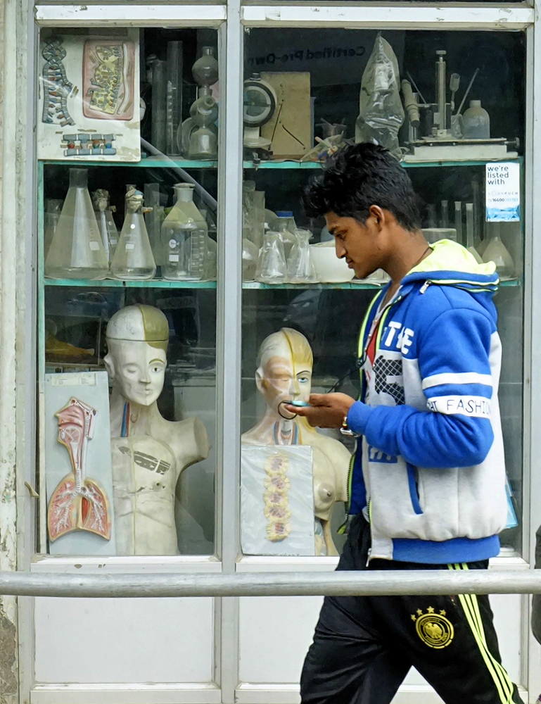 Street photography, storefront, shop window, human anatomy, anatomical model, laboratory glassware, scientific equipment, man with cell phone, passer-by,  Nepal